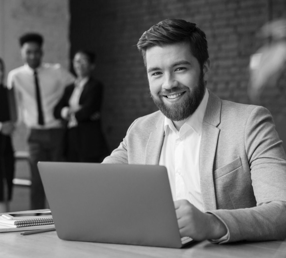 Image of cheerful young caucasian businessman sitting indoors using laptop computer. Looking camera.